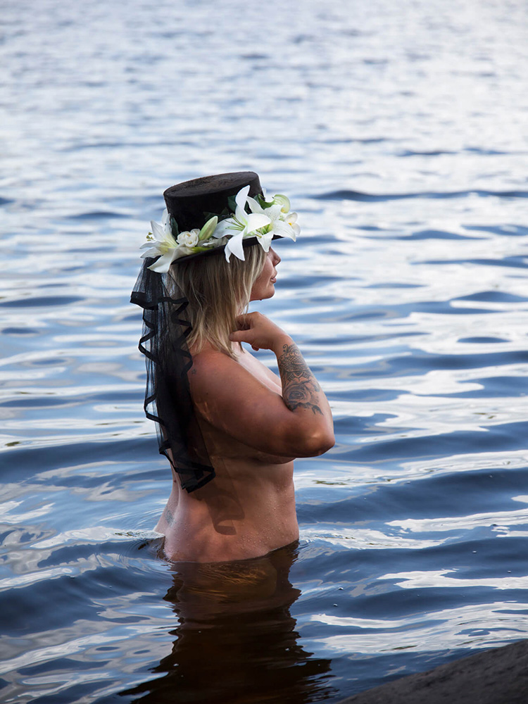 sjodrottning-svart-hatt-blommor-vatten-portratt Kvinna i sjö med svart hatt och vita blommor i ett dramatiskt vattenporträtt.