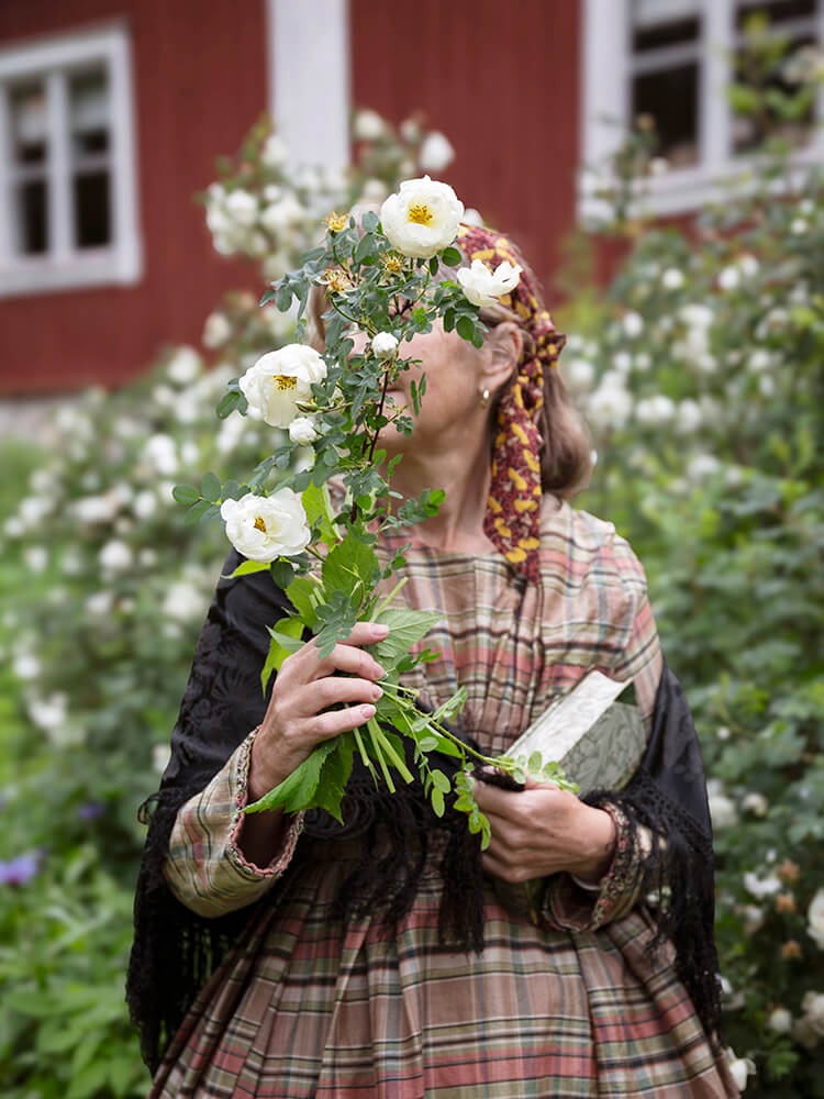 wira-kvinnor-tinghus-rosor-viktoria-davidsson Kvinna i tidstypiska kläder från tidigt 1900-tal står framför tingshuset i Wira Bruk och håller en bukett vita rosor framför ansiktet.
