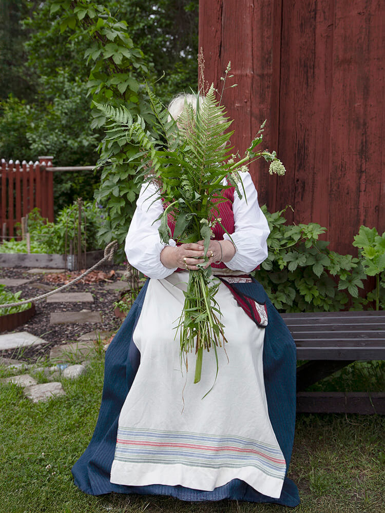 wiras-kvinnor-bank-sommarblommor-viktoria-davidsson Kvinna i folkdräkt sitter på en bänk utomhus och håller en bukett sommarblommor framför ansiktet.