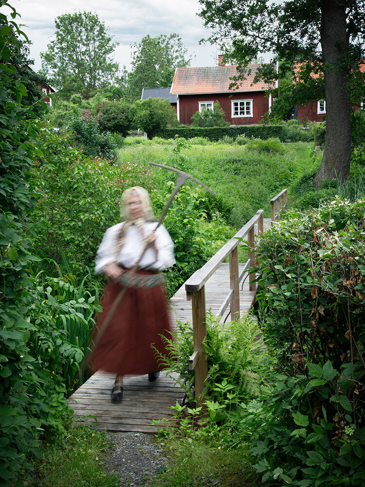 wiras-kvinnor-bro-redskap-kvinnohistoria-viktoria-davidsson En kvinna i tidstypiska kläder går över en bro och håller ett avlångt redskap.