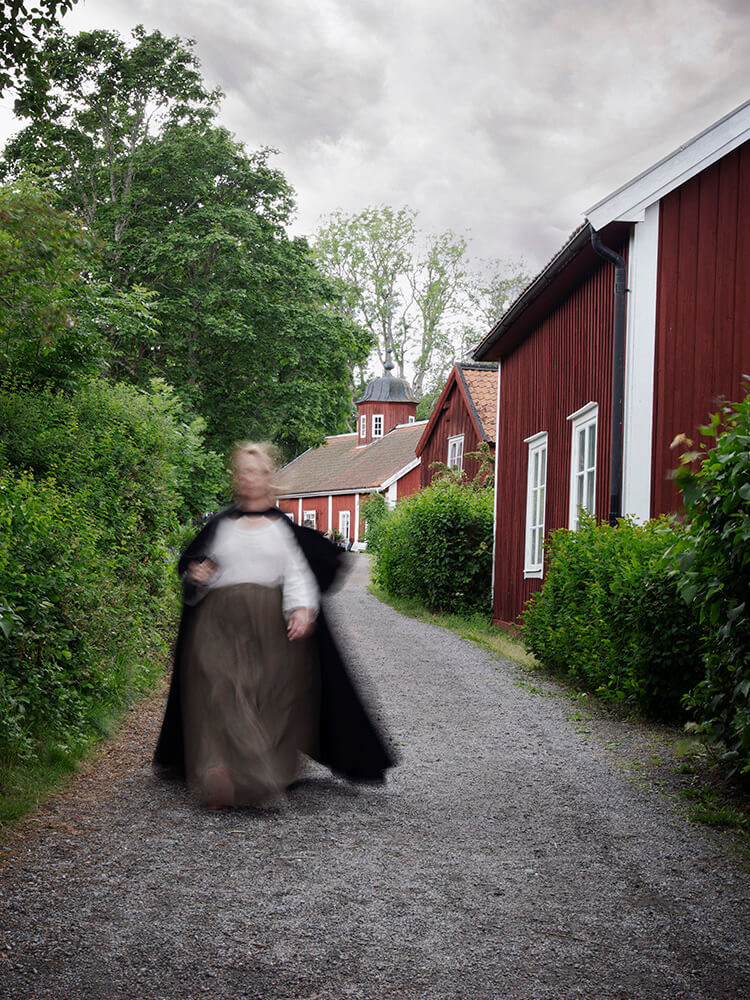 wiras-kvinnor-bruket-kvinnoportratt-viktoria-davidsson En kvinna i tidstypiska kläder går längs en väg med rörelseoskärpa som döljer ansiktet.