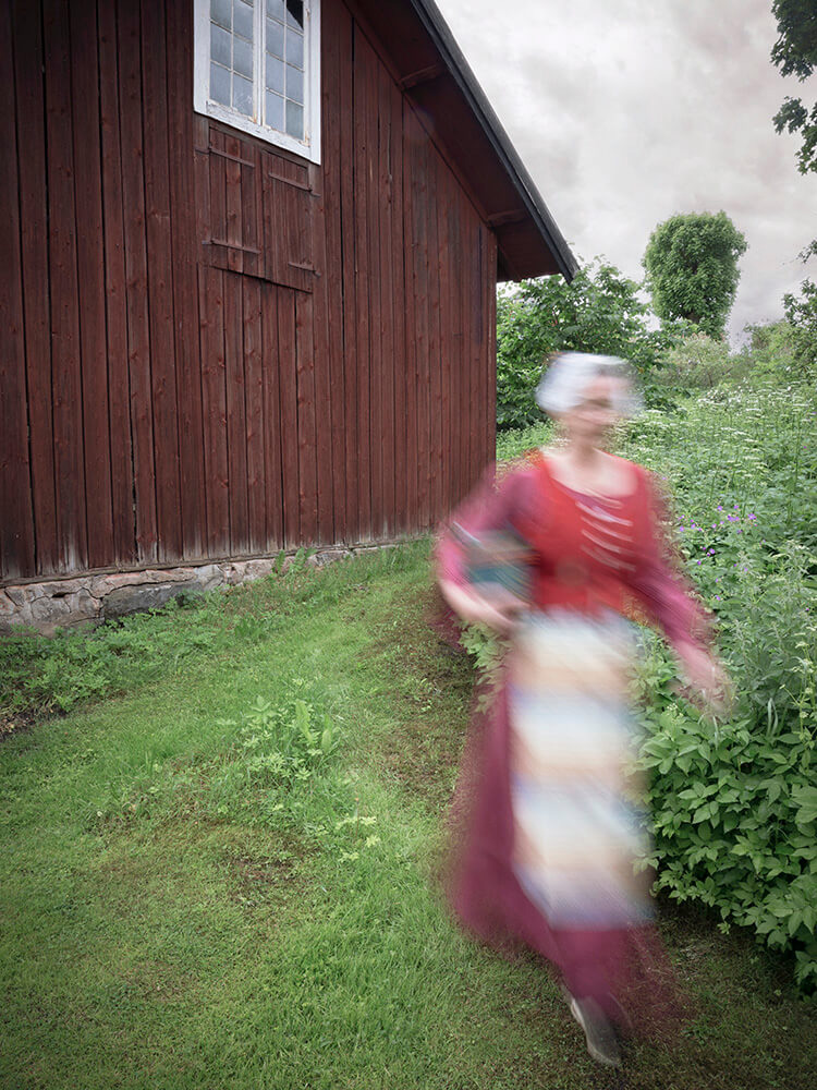 wiras-kvinnor-pa-vag-viktoria-davidsson Kvinna i tidstypiska kläder går med ett blått föremål under armen.