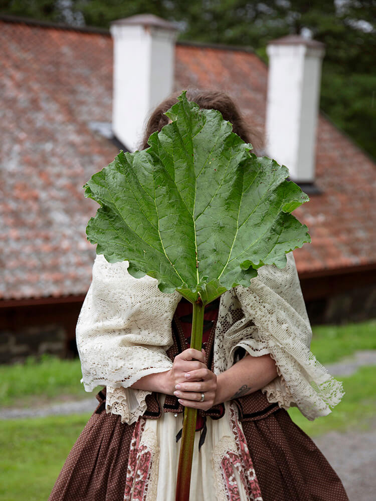 wiras-kvinnor-smedja-rabarberblad-viktoria-davidsson Kvinna i tidstypiska kläder står framför smedjan och håller ett rabarberblad framför ansiktet.